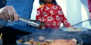 妈妈用手把猪肉、香肠、蘑菇和芦笋烤在平底锅上，用麻辣调味。亚洲家庭在松林露营时在篝火上做饭。