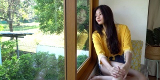 快乐年轻美丽的亚洲女人放松在房间的窗户在家里。漂亮的少女独自坐着，看着外面。肖像迷人的年轻女子脸上微笑的概念。