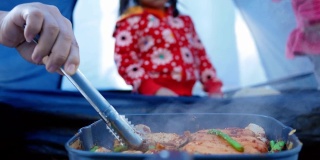 妈妈用手把猪肉、香肠、蘑菇和芦笋烤在平底锅上，用麻辣调味。亚洲家庭在松林露营时在篝火上做饭。