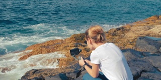 一位金发碧眼的博主在智能手机上用万向节稳定器拍摄冲浪视频。在海洋的火山海岸。为社交网络制作内容的概念
