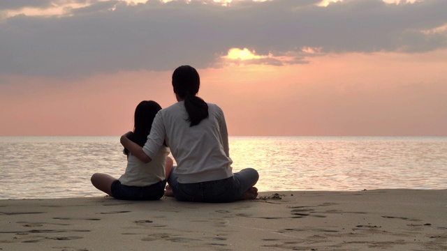 剪影快乐的母亲和女儿坐在海边的沙滩上玩日落海浪。积极的父母和人在暑假与孩子户外活动。幸福的家庭假期。家庭、生活方式、人、人寿保险、假期