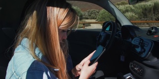 一位穿着牛仔夹克的金发美女坐在汽车副驾驶座上，用智能手机在社交网络上发送信息。阳光照亮了模特儿的头发