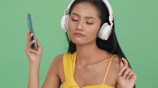 亚洲美女听音乐在线和幸福的情绪微笑。女人戴着耳机，拿着智能手机。美的概念，时尚和音乐体验。
