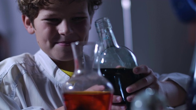 特写脸的高加索男孩混合黑色和红色化学试剂。穿着实验服的少年拿着装满各色液体的烧瓶。智能，化学，z世代电影4k ProRes HQ。