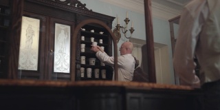 高级白种药剂师从货架上拿起装有药品的老式容器，与顾客交谈。19世纪的药店，复古，古董，医药博物馆。