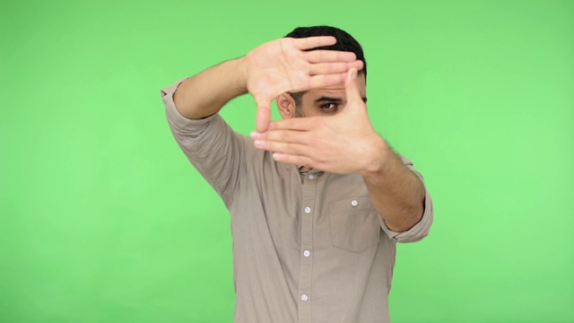 黑发男子用手指透过镜框形状看，用专业摄影师的目光盯着相机，捕捉照片。绿色背景，色度键