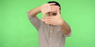 黑发男子用手指透过镜框形状看，用专业摄影师的目光盯着相机，捕捉照片。绿色背景，色度键