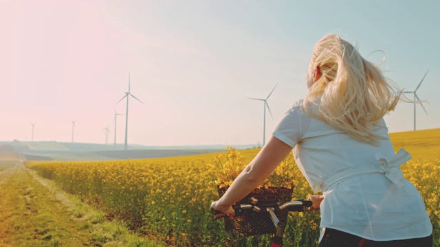 女人骑着自行车沿着油菜籽田走，远处有风力发电机