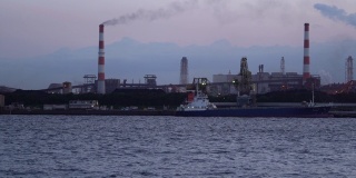 一艘工业船在燃煤电厂前的码头卸煤，烟囱里冒出滚滚浓烟。