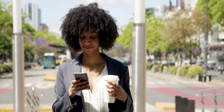 美丽的黑色商务女性在智能手机上发短信，同时享受一个外卖咖啡看起来深思熟虑