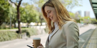 年轻漂亮的女商人用她的智能手机发短信等公交车，享受着外卖咖啡