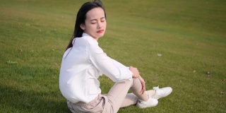 美丽的中国年轻女子坐在绿色的草地上，闭着眼睛，享受微风吹拂她的长发，最后睁开眼睛微笑，4k视频，慢镜头。