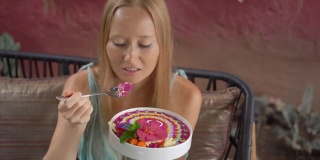 年轻女子坐在美丽的咖啡馆里，享受着盛在碗里的彩色麦片奶昔。健康饮食的概念。Slowmotion拍摄