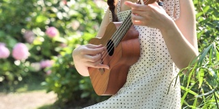 一个穿着裙子的女人的手拿着和弹奏四弦琴在花园与模糊的公园背景，4k视频，慢动作。