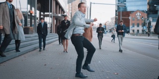 一名身穿灰色西装的兴高采烈的年轻商人正在城市街道上翩翩起舞。办公室经理穿过繁忙的办公室人群通勤上班。以摩天大楼为背景的市中心。