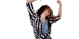 年轻女子在格子衬衫听音乐使用耳机站在孤立的白色背景非常高兴和兴奋做手臂举起的舞蹈，情绪感动。慢动作