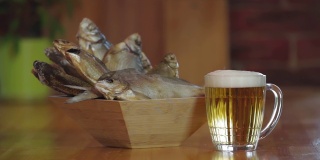 一只手拿起一杯啤酒。啤酒和鱼干。木质吧台上放着一杯清凉的淡啤酒。小吃,啤酒。装干鱼的木碗。