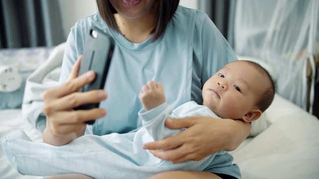 亚洲年轻母亲和男婴坐在床上，用智能手机进行视频通话