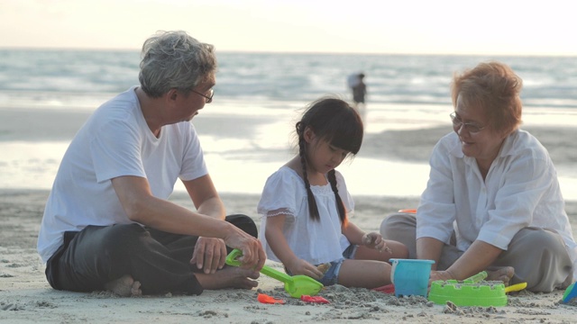 快乐的爷爷、奶奶和孙女在沙滩上的沙箱里为孩子们玩玩具。家庭，生活方式，人，老人，人寿保险，假期，关系，假期，孩子，退休，健康保健理念。