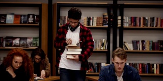 一个穿着格子衬衫戴着耳机的家伙带着一堆书走在旁边的慢动作镜头。其他同学坐着，在学院、大学图书馆里学习。前视图