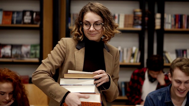 一个穿着棕色夹克，戴着眼镜的女孩拿着书架上的一堆书走在路上的慢镜头。其他同学坐着，在学院、大学图书馆里学习。前视图