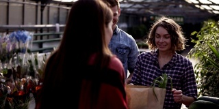 一对欧洲年轻夫妇收到的有机食品在棕色纸袋从女卖家在温室。一名男子用美元支付。笑容可掬的妇女在餐桌上出售有机食品