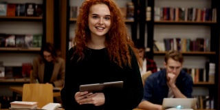 这是一幅迷人的长红头发欧洲女学生的肖像，她站在高中图书馆里，微笑着看着相机，手里拿着平板电脑。教育、文学和人的观念。背景模糊的同学
