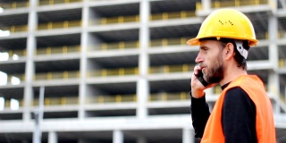 工程师或工人戴着白色头盔在模糊的建筑工地背景前打电话。