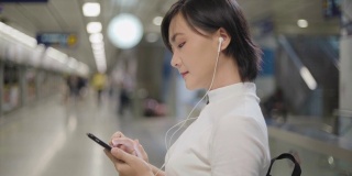 亚洲女性用耳机听音乐，用智能手机与朋友聊天或在等火车时浏览网页。日常生活和旅行中的科技。