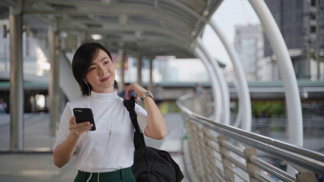 亚洲妇女的肖像用耳机听音乐和使用智能手机聊天或浏览沿着城市的街道。日常生活和旅行中的科技。