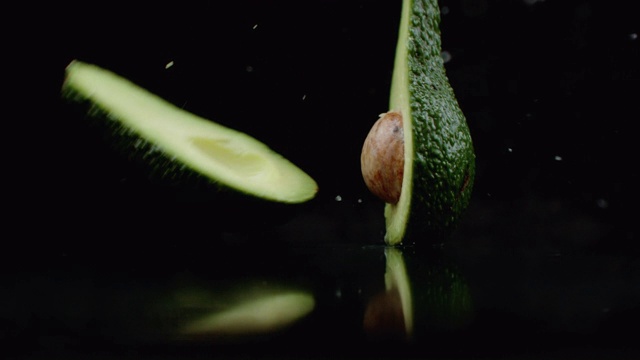 一个熟透的绿色鳄梨掉落在一个玻璃杯上，水在黑暗的背景上以慢镜头溅落。