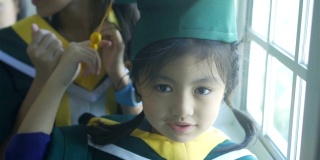 亚洲小学女生戴着毕业帽，穿着毕业服，站在学校里微笑着。