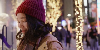 一个亚洲女孩站在商店的橱窗外在一个寒冷的夜晚在城市的圣诞市场，她穿着暖和的衣服。