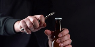 男人打开冰啤酒瓶盖上的黑色背景。用开瓶器在黑色背景上用手打开冷藏的小麦或啤酒