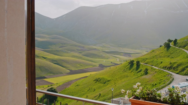 女士打开酒店的窗帘，揭示阳光明媚的山景，Castelluccio，翁布里亚，意大利