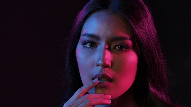 近距离肖像拍摄的年轻调情可爱的亚洲女人调皮摆姿势的相机在粉红和蓝色霓虹灯在慢动作。