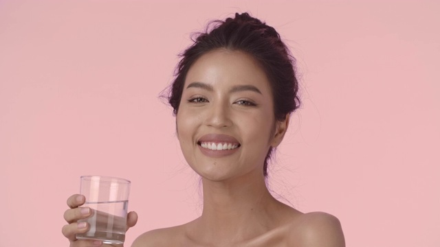 迷人的亚洲女人喝水。特写美丽的年轻亚洲女人与完美的光滑健康的皮肤喝水从一个杯子和微笑看着相机在粉红色的背景。
