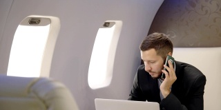 英俊的首席银行家董事商人正在打电话，工作与笔记本电脑坐在飞机内部