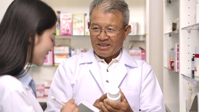 两名亚洲药剂师在药房检查药品。高级男药师和他年轻的女同事站在药房的货架前拿着药，用触控式电脑互相咨询。