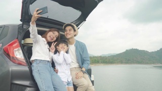 亚洲家庭坐在汽车与父亲，母亲和女儿自拍与湖泊和山景通过智能手机在假期一起度假。幸福的家庭时间。视频素材模板下载