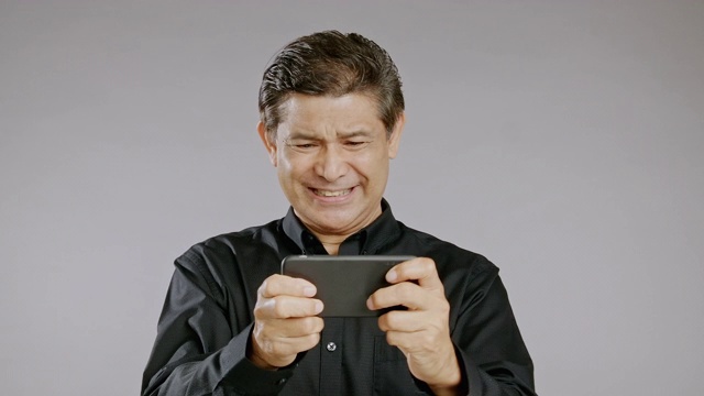 老人玩着自己的手机游戏，微笑着，大获全胜，孤立在灰色背景中。穿着黑衬衫的亚洲人