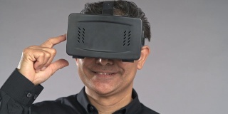 近景老人使用虚拟现实耳机，按下按钮，微笑，孤立在灰色背景中。穿着黑衬衫的亚洲人。