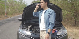 亚洲年轻的不快乐的男人在打开的引擎盖坏了的汽车前打电话在乡村路打电话寻求帮助。在旅途中，路上的破车。