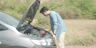 亚洲年轻的不高兴的男人在打开的引擎盖前检查坏了的汽车引擎坏了的汽车在乡村路上等待道路援助服务。在旅途中，路上的破车。