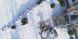 一场冬季降雪后，一名50岁的长头发、身穿黄色夹克的男子用铁铲在乡村房屋前院的小路上清理积雪。