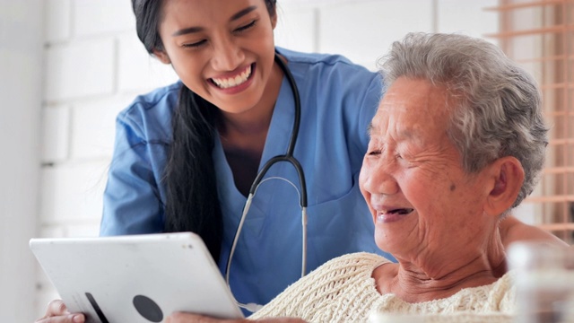 亚洲老年妇女与年轻的黑人女护士医生交谈，帮助患者在家里进行平板电脑治疗Covid-19病毒的教育。教育、医疗、护理、技术、老年护理、退休、志愿者、慈善、养老院、冠状病毒或新冠肺炎