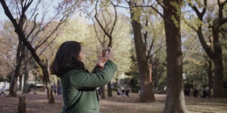亚洲年轻女游客享受公共公园的氛围，并从手机拍照。她穿着暖和的衣服。