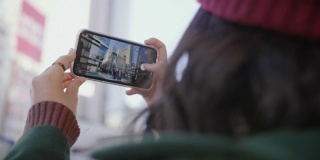 亚洲女孩喜欢这个城市，在公交车站等车的时候用智能手机拍照或录像。