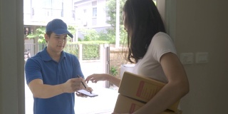亚洲快递年轻男子在蓝色制服微笑，抱着一堆纸箱在前面的房子和亚洲妇女接受从快递员送的盒子。广告，商业，交通概念。
