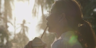 年轻的亚洲女人喝椰子水在日落，阳光周末公园